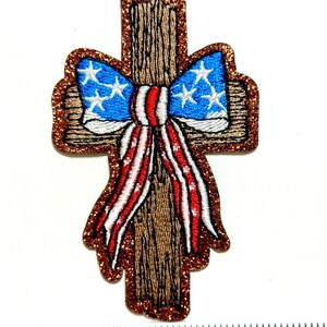May include: Embroidered patch in the shape of a cross with a bow. The bow features a blue background with white stars and red and white stripes. The cross has a brown, wood-like texture, and the border is glittery.