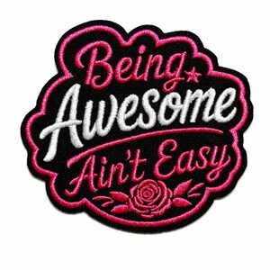 May include: An embroidered patch with a black background and a pink border. The text reads Being Awesome in white and Ain't Easy in pink. A small pink star is next to the word Awesome, and a pink rose with leaves is at the bottom center.