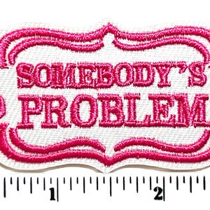 May include: Embroidered patch with the phrase "SOMEBODY'S PROBLEM" in pink lettering. The text is inside a white rectangular shape with a pink decorative border. The patch is approximately 3 inches wide.