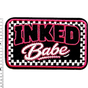 May include: A rectangular embroidered patch with a black center, white and black checkered border, and a pink outer edge. The text INKED in black with pink outline and Babe in white script is centered. A ruler shows a width of 2 inches.