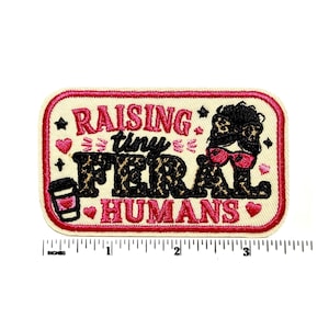 May include: A rectangular embroidered patch with a cream background and a pink border. It features the text "RAISING tiny FERAL HUMANS" in pink and leopard-print black lettering, a coffee cup, and a messy bun icon. A ruler shows it is 3 inches wide.