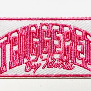 May include: Embroidered patch with the words "TRIGGERED" in a pink, stylized font, arched across the top. Below, in smaller pink script, it reads "By Idiots." The patch is rectangular with a white background and a pink border.