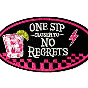 May include: Oval-shaped black patch with a pink border and the text "ONE SIP CLOSER TO NO REGRETS" in white. A pink cocktail with ice and a lime wedge is on the left. A pink lightning bolt is on the right. The patch is approximately 2 inches long.
