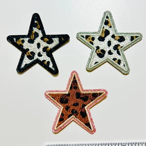 May include: Three star-shaped patches with a leopard print design. One patch has a black border, another has a silver border, and the third has a pink border. The leopard print features black and brown spots on a white background.