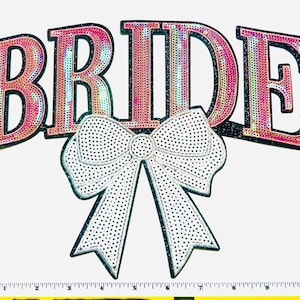 May include: A decorative sign with the word "BRIDE" in pink, glittery letters with a black outline. A white bow with black polka dots is centered below the word. The sign is likely for a wedding or bridal event.