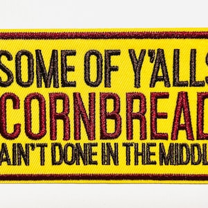 May include: Yellow embroidered patch with a brown border and text that reads "SOME OF Y'ALLS CORNBREAD AIN'T DONE IN THE MIDDLE" in brown and red thread. The patch is rectangular.