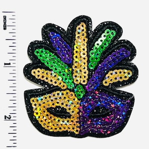 May include: A sequined Mardi Gras mask patch in black, gold, green, and purple. The mask is approximately 2 inches tall and features a glittery black border. The sequins are arranged in a decorative pattern.