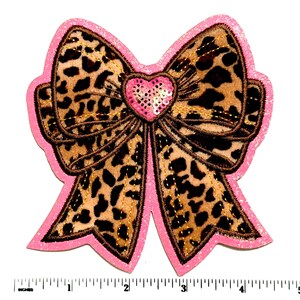 May include: A bow-shaped patch featuring a tan and black leopard print pattern with a pink glitter border. A small pink heart with sequins sits at the center. A white measuring tape at the bottom shows the patch is approximately 5.5 inches wide.