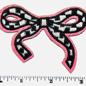 May include: A decorative bow-shaped patch with a pink glitter border. The bow features a black and white checkered pattern. The patch is approximately 3 inches wide.