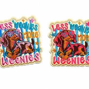 May include: Two identical embroidered patches featuring a brown dachshund wearing heart-shaped glasses. The text reads Less meanies more weenies in pink and orange. The background has blue vertical stripes and small colorful stars.