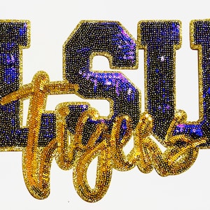 May include: A sequined patch featuring the letters "LSU" in blue with gold trim, and the word "Tigers" in gold script. The patch is approximately 5 inches wide and 4 inches tall, with a white background.