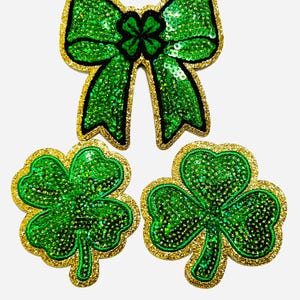Puede incluir: Tres parches temáticos del Día de San Patricio. Un lazo verde con lentejuelas con un borde dorado brillante y un contorno negro se encuentra encima de dos parches de trébol verde con lentejuelas con bordes dorados brillantes.