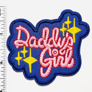 May include: A blue embroidered patch with the words "Daddy's Girl" in pink script. The patch features yellow starburst designs and a blue border. The patch measures approximately 2 inches in height.