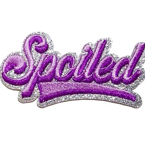 May include: A purple embroidered patch with the word "Spoiled" in a cursive font. The text is outlined in a silver glitter material. The patch measures approximately 2 inches in length.