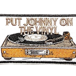 May include: Embroidered patch featuring a vintage-style record player. The patch is rectangular with a black border and the text "PUT JOHNNY ON THE VINYL" in gold thread. The record player is detailed in shades of brown, black, and gold.