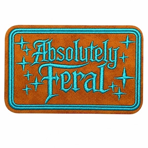 May include: A rectangular embroidered patch with a brown felt-like background and a bright turquoise border. The text Absolutely Feral is stitched in turquoise thread in a stylized font, surrounded by several small four-pointed star shapes.