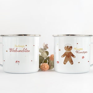 May include: Two white enamel mugs with festive Christmas designs. One mug features the text "Frohe Weihnachten" in red lettering with gold stars and hearts. The other mug features a gingerbread man with the text "Kommer Kommer" in red lettering with gold stars and hearts.