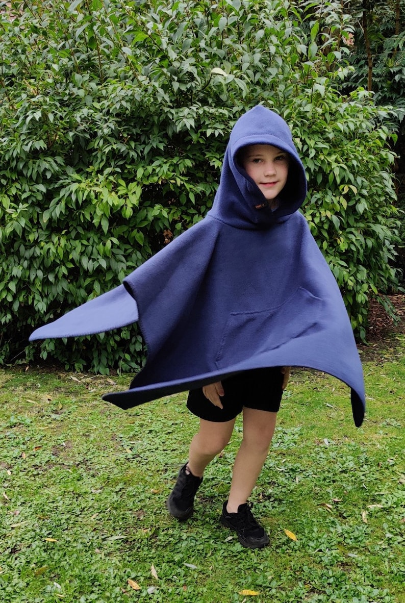May include: A child wearing a navy blue hooded poncho with a pocket. The poncho is made of fleece and has a drawstring hood. The child is standing in a grassy area.