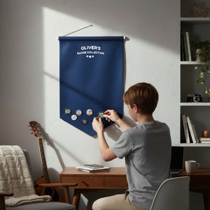 May include: A dark blue fabric wall banner with a white wooden dowel and hanging string. The text OLIVER'S BADGE COLLECTION is printed in white at the top, accompanied by three small stars. Several circular pin-back badges are attached to the fabric.