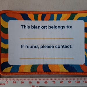 May include: Rectangular blanket tag with an orange border and a colourful wave pattern. The tag has the text "This blanket belongs to:" and "If found, please contact:" with lines for writing. A ruler is at the bottom.
