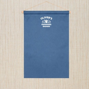 May include: A blue fabric wall banner suspended from a wooden dowel with a string. The banner features a white graphic of a person swimming above the text "OLIVER'S SWIMMING BADGES" printed in white capital letters in the upper centre section.