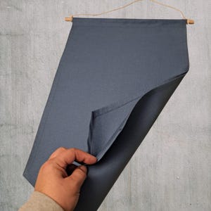 May include: A slate gray wall hanging with a wooden dowel and twine for suspension. The fabric is folded to reveal the underside, showcasing a clean, finished edge. The background is a textured gray wall.