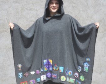 May include: A grey hooded poncho with a variety of colourful patches sewn on the bottom edge. The patches feature different designs, including a snowflake, a compass, a flower, and a bird.