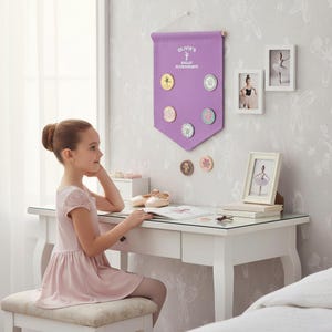 May include: A lilac banner with the text "OLIVIA'S BALLET ACHIEVEMENTS" displays ballet achievement medallions. The room includes a white desk, ballet shoes, and framed ballet photos. A young girl in a pink dress sits at the desk, with a ballet theme.