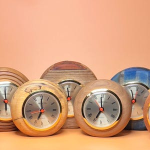 May include: A collection of wooden sphere-shaped clocks with silver faces and red hands. The clocks vary in wood grain and color, including natural, blue, and brown tones. The clocks have a gold-colored bezel.