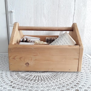 Könnte beinhalten: Eine hellfarbene Holzwerkzeugkiste mit Griff, die kleine Gegenstände wie Spitze, ein Foto und eine handschriftliche Notiz enthält. Die Kiste steht auf einer weißen, gemusterten Oberfläche vor einem weißen Holzhintergrund.