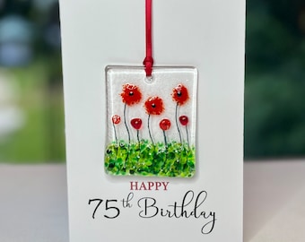 May include: A white card with a red ribbon attached to a glass ornament featuring red poppies with green leaves. The card reads "Happy 75th Birthday".