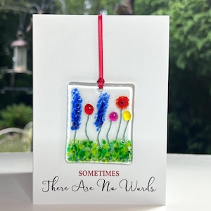 May include: A white card with the text "Sometimes There Are No Words" printed on it. A square glass ornament with a red ribbon is hanging from the card. The ornament features a colourful glass flower design with blue, red, yellow, and pink flowers.
