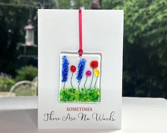 May include: A white card with the text "Sometimes There Are No Words" printed on it. A square glass ornament with a red ribbon is hanging from the card. The ornament features a colorful glass flower design with blue, red, yellow, and pink flowers.