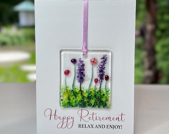 May include: A white card with a pink ribbon attached to a square glass ornament. The ornament features a purple flower with green leaves and red dots. The card reads "Happy Retirement Relax and Enjoy!"