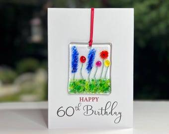May include: A white card with a red ribbon and a glass ornament featuring colorful flowers. The card reads "Happy 60th Birthday".