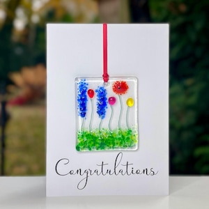 May include: A white card with the word "Congratulations" written in black cursive. A red ribbon hangs from the top of the card and is attached to a square glass ornament with a colourful floral design. The ornament features blue, red, yellow, and green glass flowers.