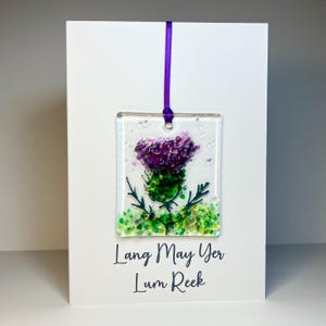 May include: A rectangular glass ornament depicting a purple thistle flower with green foliage, suspended by a purple ribbon. The ornament is set against a white card, with the text "Lang May Yer Lum Reek" displayed below the image.