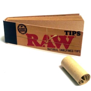 Puede incluir: Un paquete de RAW Natural Unrefined Tips, una marca de puntas de papel para enrollar. El paquete es marrón con un logotipo RAW rojo y el texto "TIPS" y "NATURAL UNREFINED TIPS". Se muestra una sola punta enrollada.
