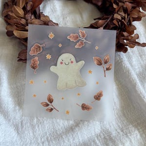 Könnte beinhalten: Ein quadratisches, durchscheinendes Pergamentblatt mit einer zentralen Illustration eines lächelnden, blassen Geistes mit rosa Wangen. Der Geist ist von kleinen, braunen Herbstblättern und winzigen gelben Sternenakzenten umgeben.