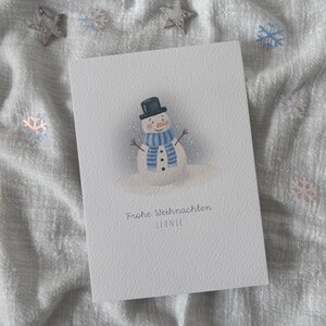 May include: A white greeting card featuring a snowman illustration. The snowman wears a black top hat and a blue and white striped scarf. The card has the text "Frohe Weihnachten LEONIE". Decorative stars and snowflakes are scattered around the card.