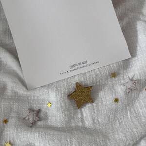 May include: A white card with the text "FOX AND THE WOLF" and "Etsy @ FoxandtheWolfCreates" is surrounded by gold glitter stars and star-shaped wooden cutouts on a white fabric surface.