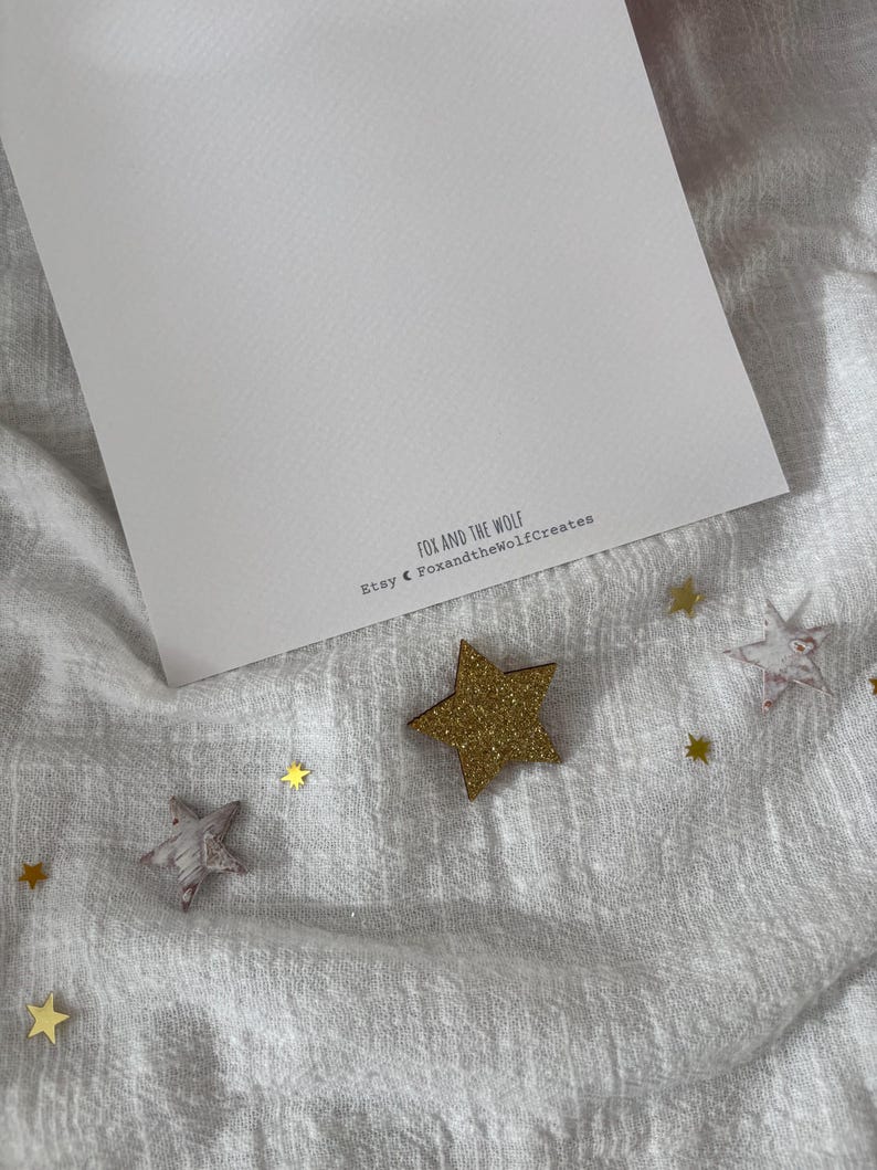 May include: A white card with the text "FOX AND THE WOLF" and "Etsy @ FoxandtheWolfCreates" is surrounded by gold glitter stars and star-shaped wooden cutouts on a white fabric surface.