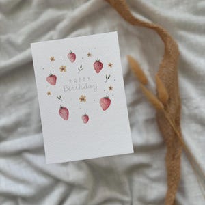 May include: A white card with the words "Happy Birthday" written in gray script. The card is decorated with a circle of red and pink strawberries, green leaves, and yellow flowers.
