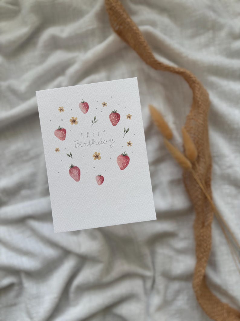 May include: A white card with the words "Happy Birthday" written in gray script. The card is decorated with a circle of red and pink strawberries, green leaves, and yellow flowers.
