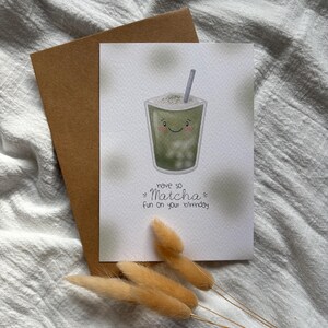 Puede incluir: Tarjeta de felicitaci&oacute;n de cumplea&ntilde;os con una ilustraci&oacute;n de bebida matcha sonriente, una pajita y el texto "have so Matcha fun on your birthday". La tarjeta va acompa&ntilde;ada de un sobre marr&oacute;n y flores secas.