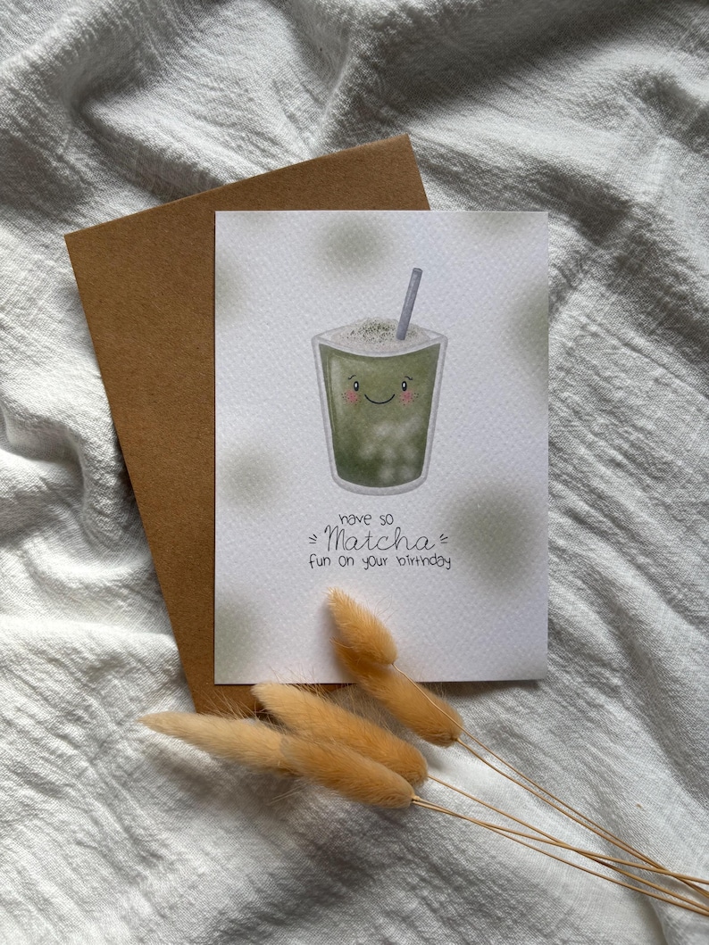 Puede incluir: Tarjeta de felicitaci&oacute;n de cumplea&ntilde;os con una ilustraci&oacute;n de bebida matcha sonriente, una pajita y el texto "have so Matcha fun on your birthday". La tarjeta va acompa&ntilde;ada de un sobre marr&oacute;n y flores secas.