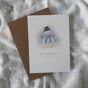 May include: A Christmas card featuring a watercolor snowman wearing a black top hat and blue scarf. The card has the text "Frohe Weihnachten Leonie" and is paired with a brown envelope. Decorative snowflakes and stars surround the card.