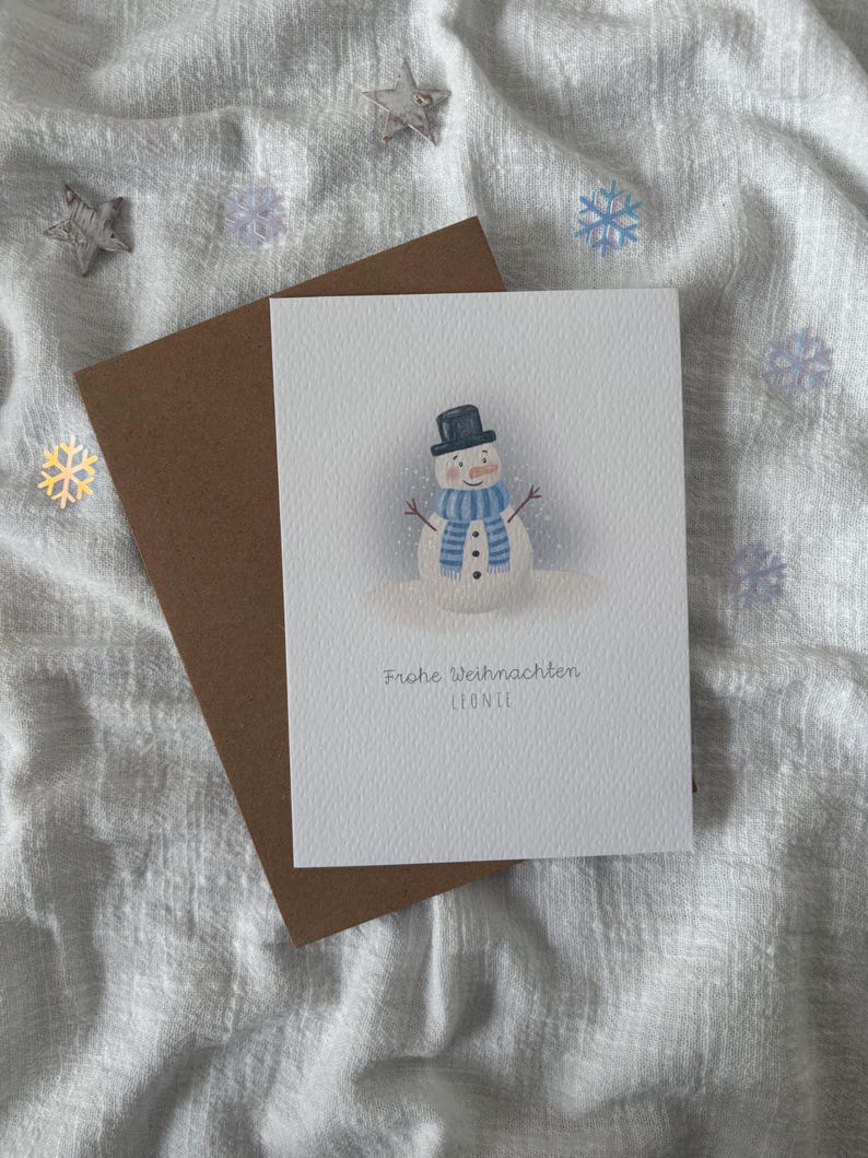 May include: A Christmas card featuring a watercolor snowman wearing a black top hat and blue scarf. The card has the text "Frohe Weihnachten Leonie" and is paired with a brown envelope. Decorative snowflakes and stars surround the card.