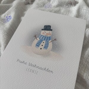 May include: A white greeting card featuring a snowman illustration. The snowman wears a black top hat, a blue and white striped scarf, and has a carrot nose. The card includes the text "Frohe Weihnachten LEONIE".