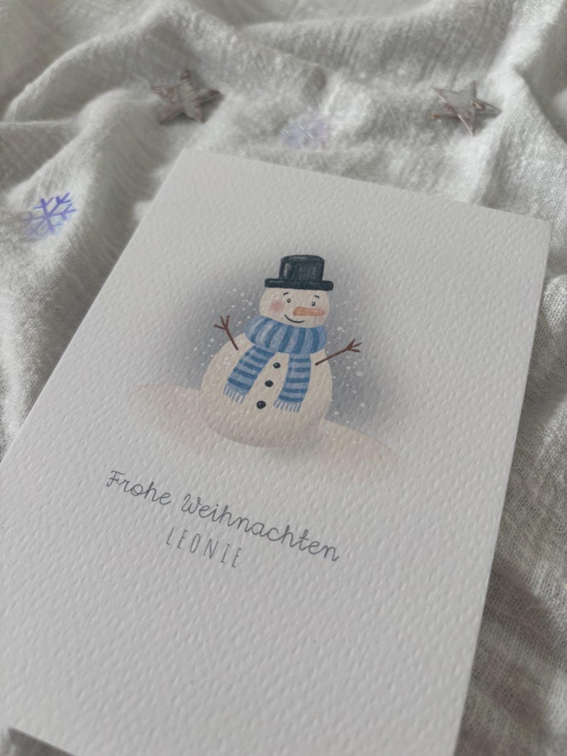 May include: A white greeting card featuring a snowman illustration. The snowman wears a black top hat, a blue and white striped scarf, and has a carrot nose. The card includes the text "Frohe Weihnachten LEONIE".